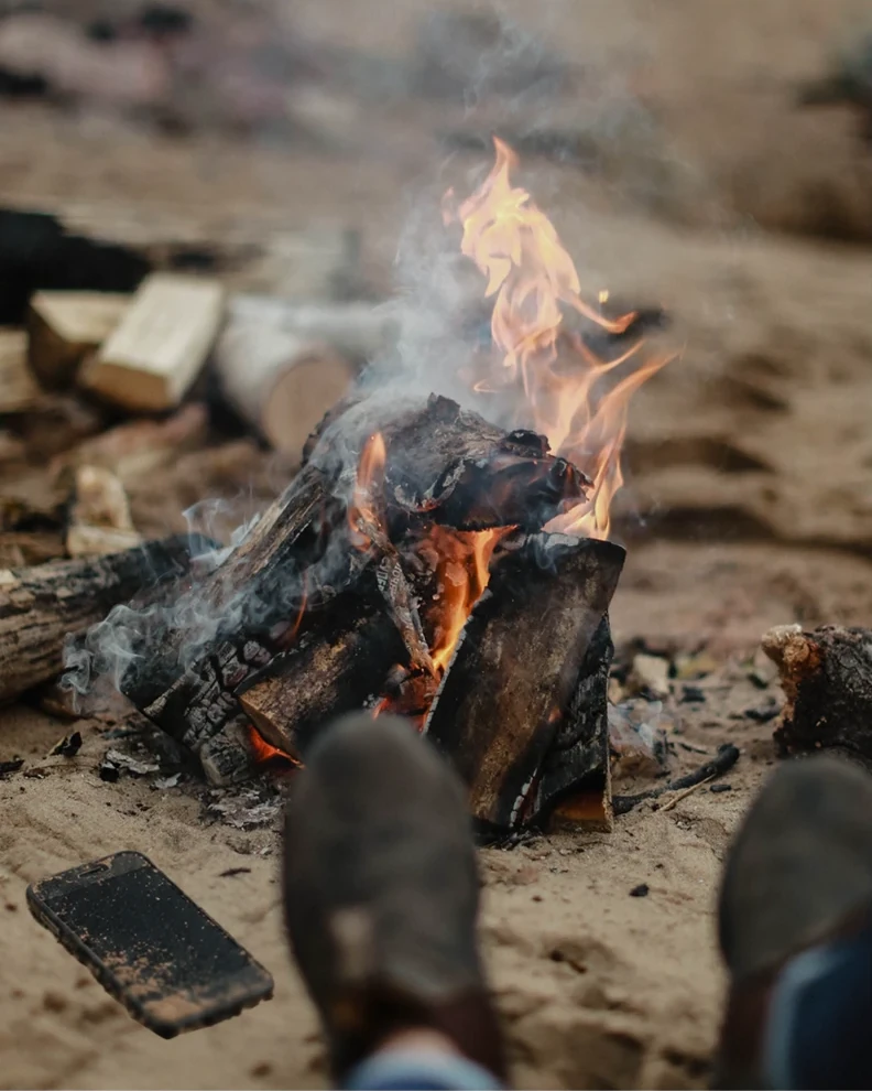 A dirty phone lying in the sand beside a campfire, someone's boots stretched out toward the flames