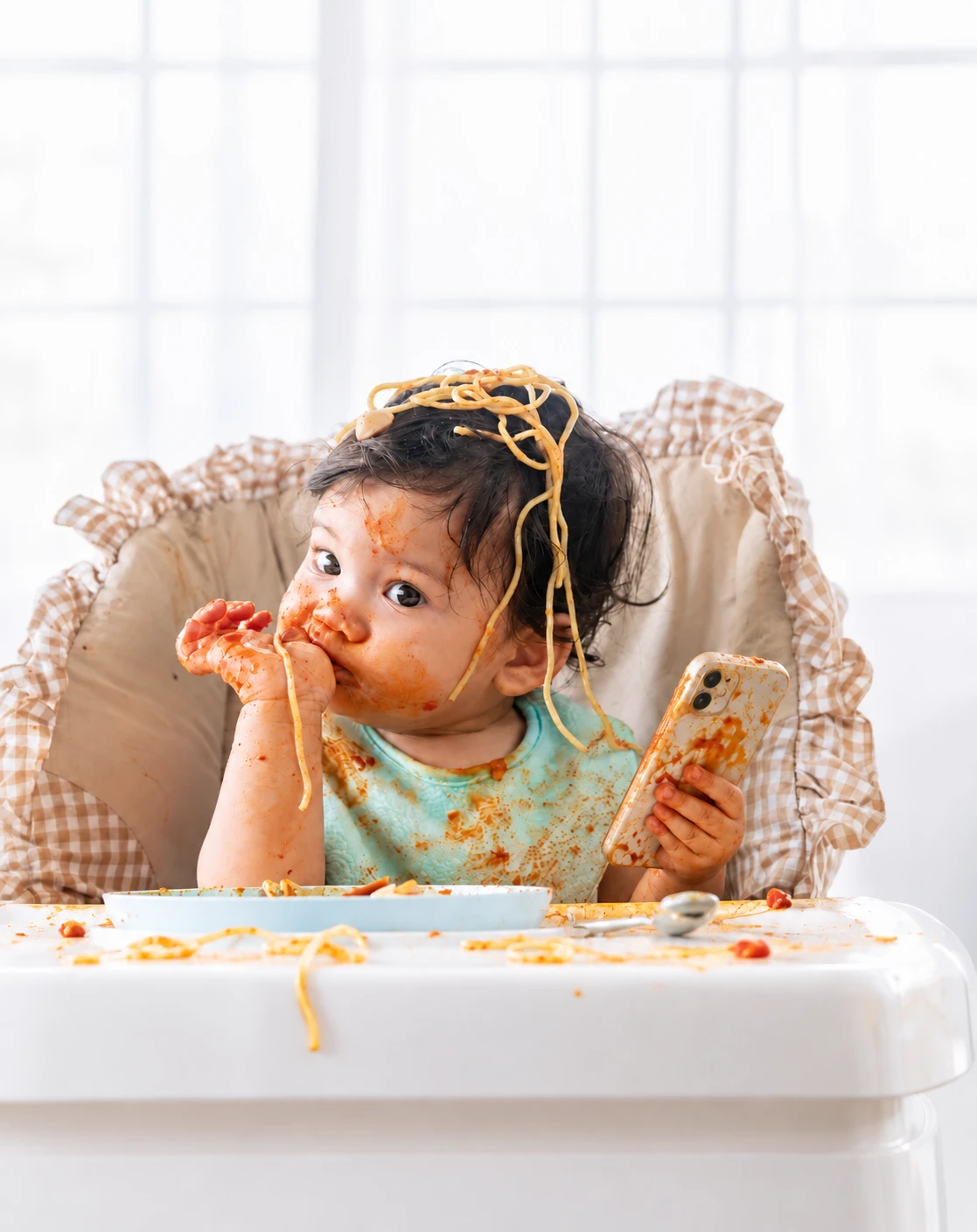 A toddler in a high chair covered in spaghetti sauce, holding a phone, noodles spilled across the tray