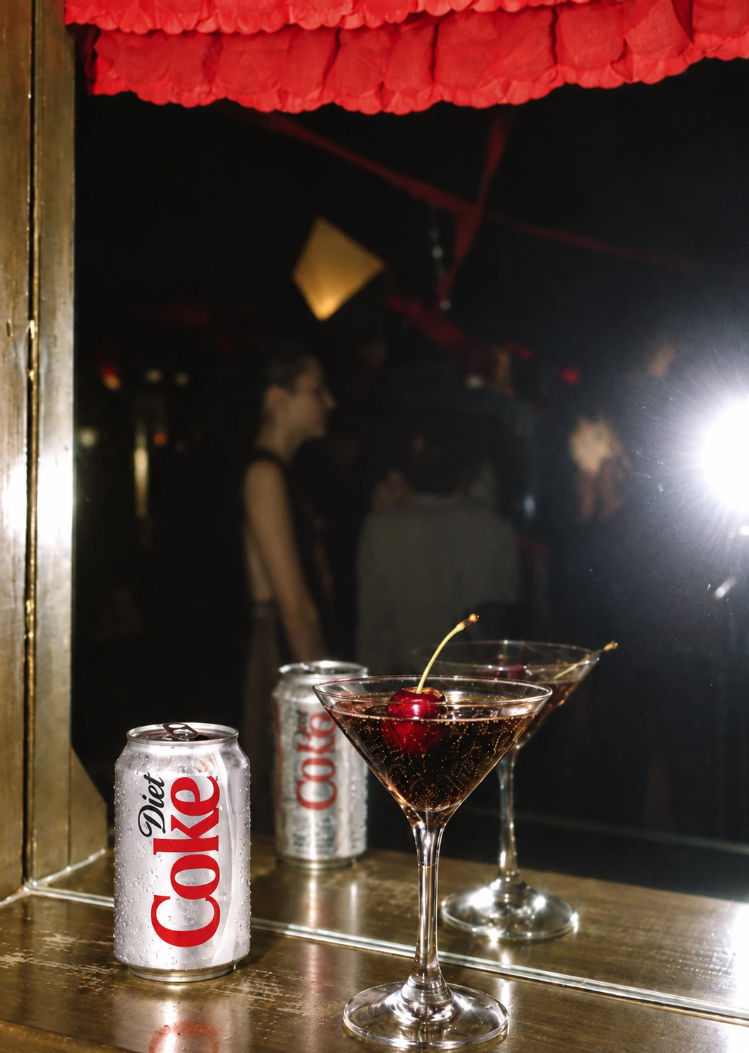 A can of Diet Coke beside a martini glass of cola with a cherry, at a dim bar
