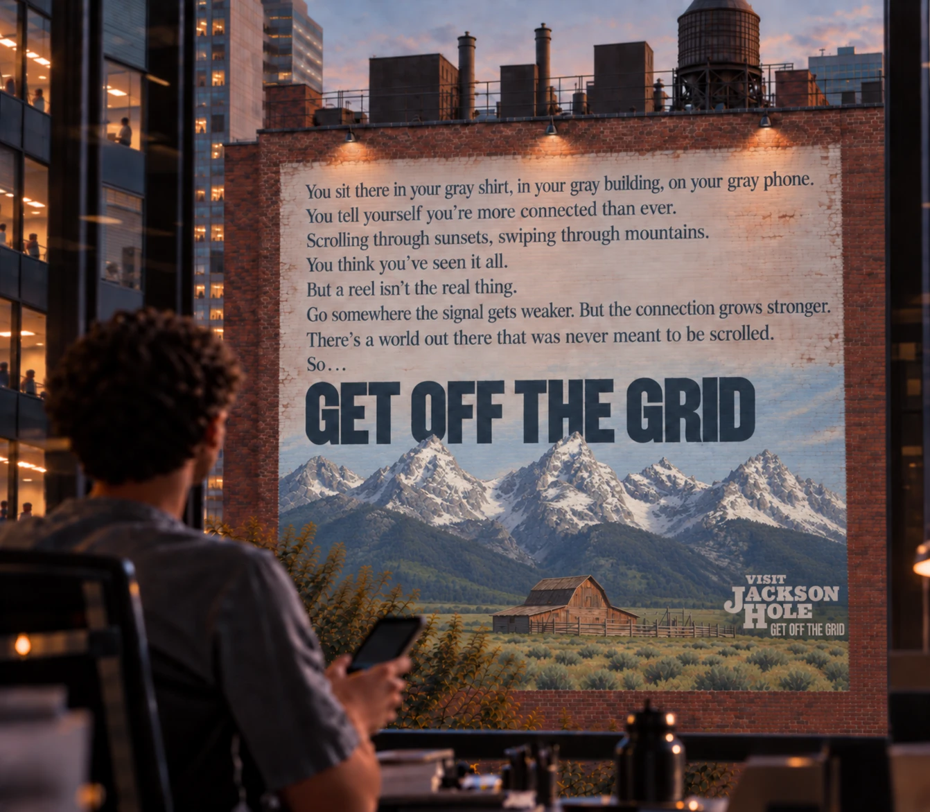 A building-side mural execution of the Visit Jackson Hole 'Get Off the Grid' campaign, seen through an office window as someone scrolls on their phone