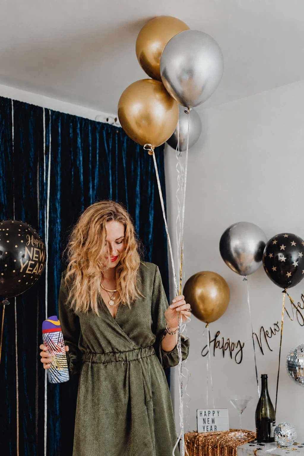 A woman in a green wrap dress holds gold and silver balloons and an Owala bottle, in front of a navy velvet curtain and Happy New Year decorations
